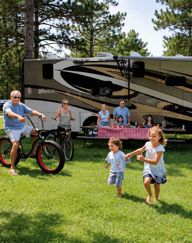 family-rv-camping-resort-membership A happy family sitting in lawn chairs outside their luxury RV camper at a private resort, watching their kids run and play on a spacious grassy campsite.