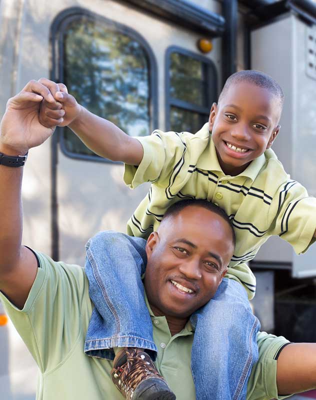 private-campground-membership-resale-savings Parents relaxing by their fifth-wheel RV trailer at a shaded private campground, watching their young children play tag on a manicured grassy lawn.